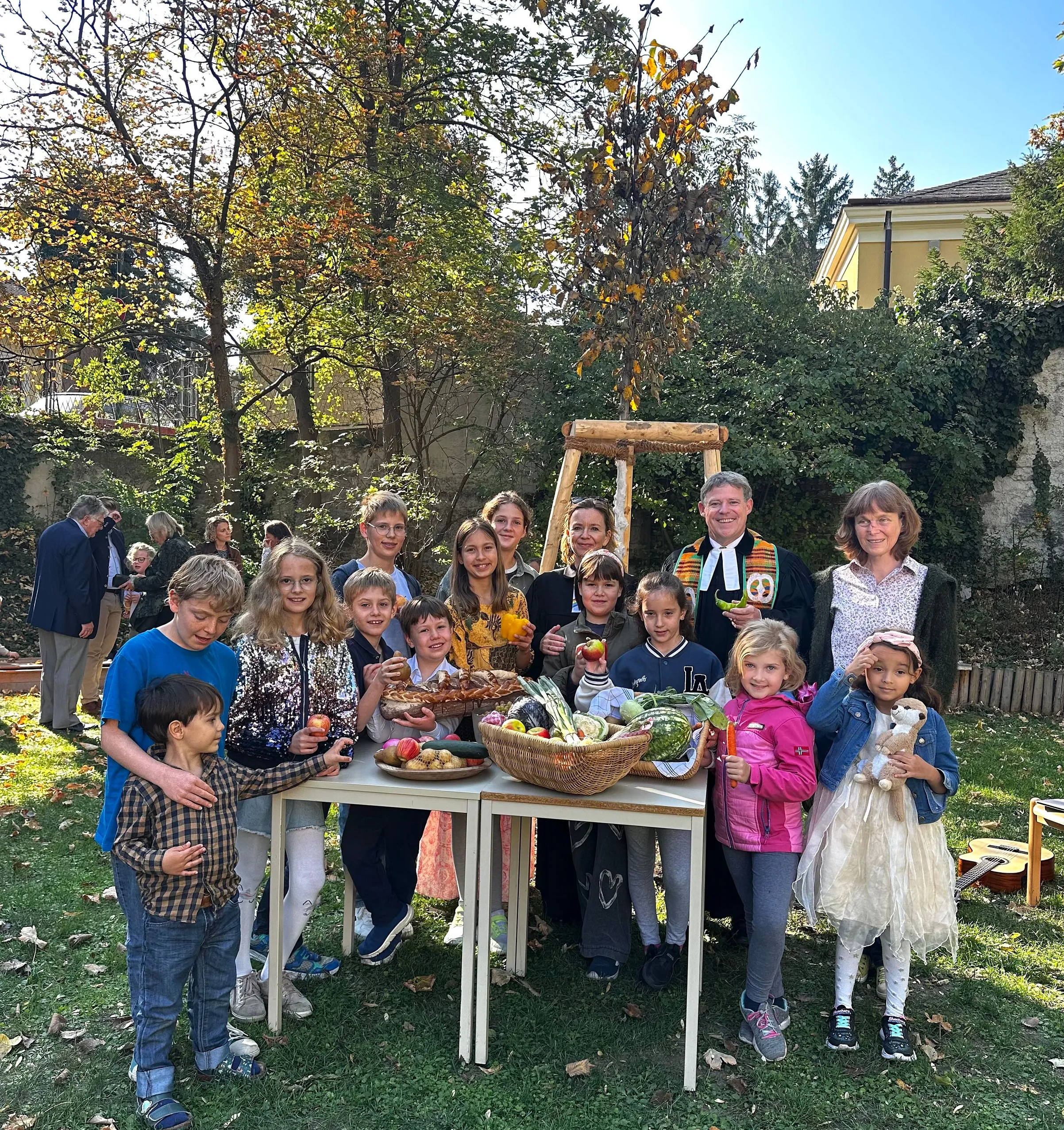 Pfarrer, Vikarin und Gemeindepädagogin und Kinder stehen in einem Garten um einen mit Obst und Gemüse gedeckten Tisch